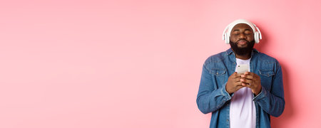 Delighted Black man enjoying awesome music, listening songs in headphones and holding smartphone, looking ecstatic, standing over pink backgroundの写真素材