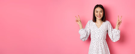 Cute asian brunette girl in dress smiling, showing kawaii peace signs and looking happy, standing over pink backgroundの写真素材