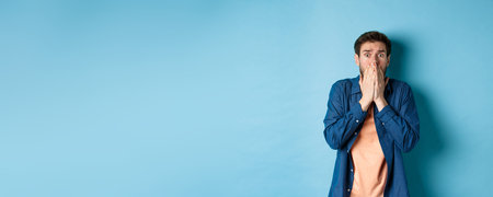 Scared anxious man covering mouth with hands and looking at something terrifying, standing on blue...
