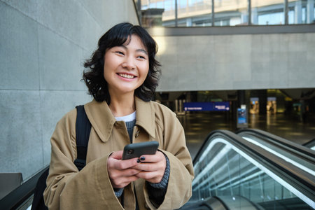 Portrait of cute korean girl in trench coat, going up escalator, holds mobile phone and smiles...