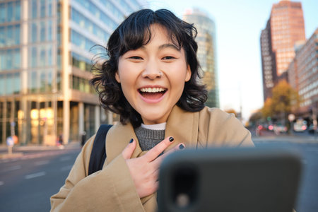Image of korean girl video chats with smartphone, looks at her phone with surprised and amazed face,...