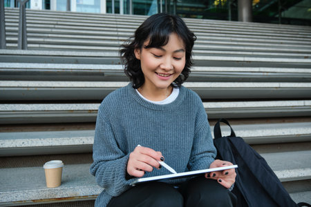 Smiling girl, graphic designer, using digital tablet and pen tool to draw, does home assignment for university, sits on stairsの写真素材