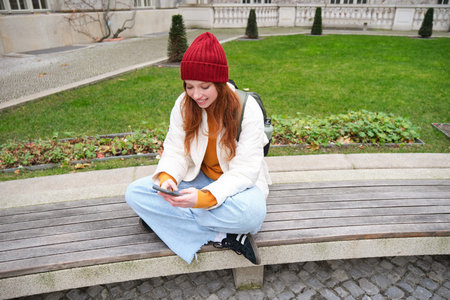 Young redhead girl student, sits on bench and uses smartphone app, watches videos online, sends message on mobile phoneの写真素材