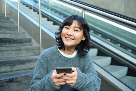 Portrait of brunette young woman, student sits on stairs in public place, looks at smartphone, reads...
