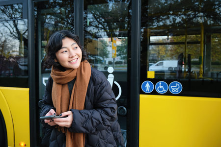 Portrait of korean girl looking for her bus on a stop, holding mobile phone, checking schedule, time...