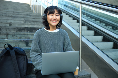 Female student works on project remotely, sits on stairs street, works on laptop, listens music in headphones and smilesの写真素材