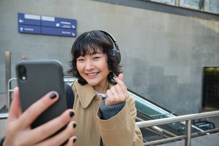 Cute and stylish Korean girl, wears headphones, takes selfie on smartphone, tourist records video or makes a photo, stands on streetの写真素材