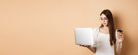 Portrait of woman working on laptop and drinking coffee. Girl student using computer for remote studies, standing on beige backgroundの写真素材