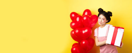 Happy Valentines day. Beautiful and stylish asian woman posing near heart balloons with lovely box with gift from lover, standing over yellow backgroundの写真素材