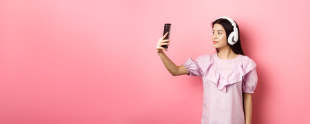 Beautiful japanese girl taking selfie on smartphone, wearing wireless headphones, standing against pink backgroundの写真素材
