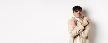 Indecisive funny young man having troubles with making choice, pointing fingers sideways and frowning perplexed, deciding between two variants, standing on white backgroundの写真素材