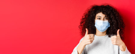 Covid-19 and pandemic concept. Close-up of young curly woman in face mask showing thumbs up, like and praise good thing, standing on red backgroundの写真素材