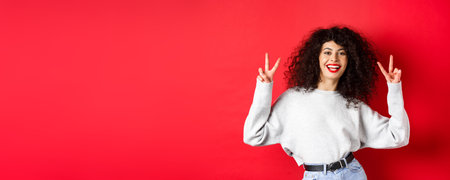 Stylish young woman with curly hairstyle, smiling happy and showing peace signs, standing in casual...