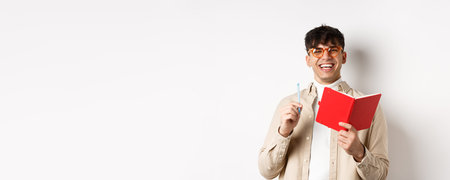 Cheerful young man in glasses laughing and taking notes, writing down in planner, holding pen and...