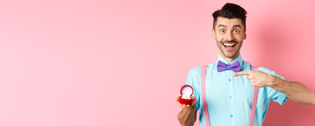 Valentines day. Cheerful young man going to make proposal, pointing finger at engagement ring and smiling excited, standing on romantic pink backgroundの写真素材
