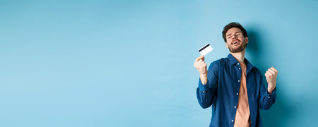 Happy and satisfied young man dancing with plastic credit card, fist pump and smile, standing on blue backgroundの写真素材