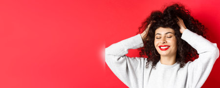 Close-up of carefree woman touching soft curly hair and smiling pleased, standing on red background. Haircare and beauty conceptの写真素材