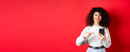 Cheerful pretty lady with smartphone pointing at her phone, smiling satisfied, recommend an app, standing against red backgroundの写真素材