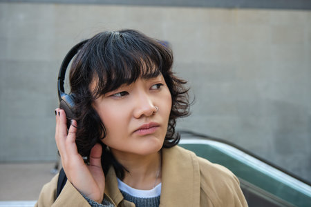 Portrait of complicated young korean girl in headphones, frowns and looks disappointed while stands...