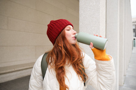 Beautiful redhead woman drinking hot tea or coffee from thermos, female tourist enjoys warm drink,...