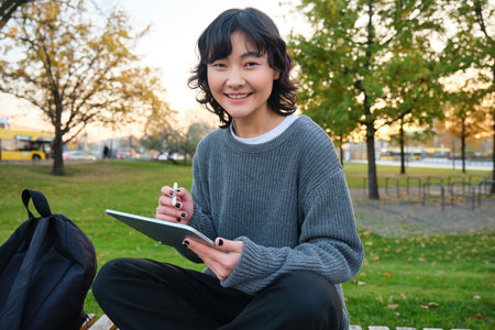 Portrait of young smiling korean girl, graphic designer, artist drawing on digital tablet with a pen tool, sitting in park on fresh air and scatching, taking notesの写真素材