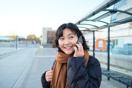 Beautiful smiling korean girl, waiting on bus stop, using public transport, talking on mobile phone, going somewhere in cityの写真素材