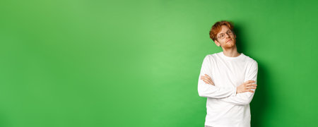 Confident young man with red hair, wearing glasses, looking smug at right side copy space, cross arms on chest self-assured, standing over green backgroundの写真素材