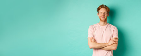 Portrait of friendly-looking young man with red hair and beard smiling and starng satisfied, holding hands crossed on chest, standing over turquoise backgroundの写真素材