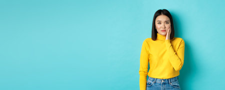 Portrait of asian young woman touching cheek and frowning, looking sad, being slapped in face, feeling painful toothache, standing over blue backgroundの写真素材