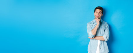 Portrait of thoughtful handsome man with beard, standing in casual outfit, looking at upper left corner and smiling, imaging or dreaming about something, standing over blue backgroundの写真素材