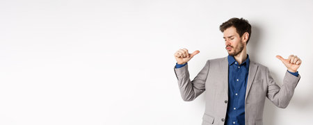 Handsome and confident man in suit pointing at himself, feeling cool and sassy, self-promoting his...