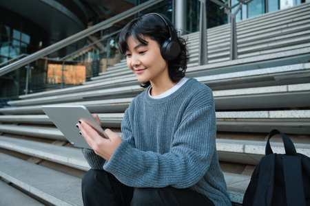 Smiling chinese student sits on stairs with tablet, draws digital art, graphic design project for...