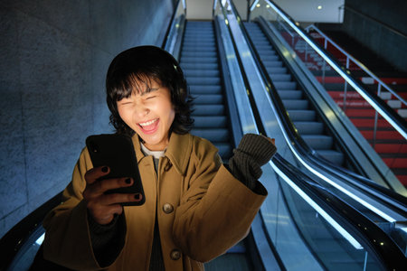 Urban lifestyle and people. Portrait of girl feeling happy and excited, going down escalator in headphones, looking at smartphone and celebratingの写真素材