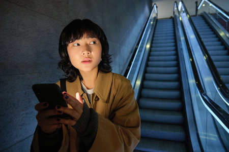 Urban lifestyle and people. Portrait of Korean girl in headphones, holds smartphone, looks concerned and tensed while going down escalator in darkの写真素材