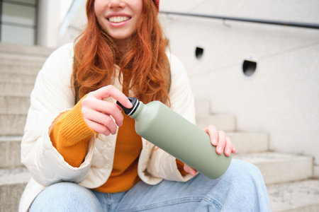 Smiling traveler, redhead girl tourist sits on stairs with flask, drinks hot coffee from thermos...