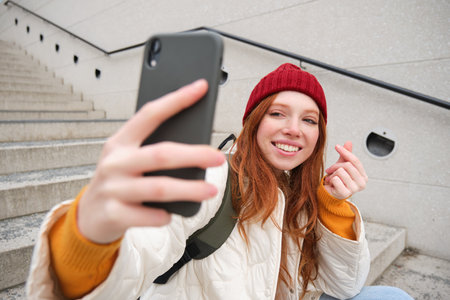 Mobile phone and people lifestyle. Stylish redhead girl takes selfie on her smartphone, poses for photo with mobile phone in hand, smiles happily, sits on stairs outdoorsの写真素材