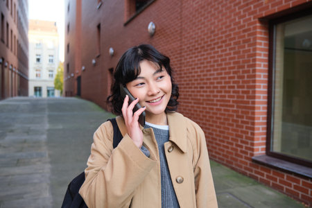 Portrait of young korean woman walking down street with phone, talking with someone, makes a call, has telephone conversationの写真素材