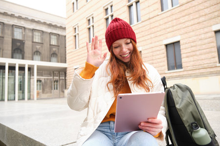 Happy redhead girl, student sits with backpack and digital tablet on street, says hello and waves at gadget camera, connects to public wifi to video chat, has online conversationの写真素材