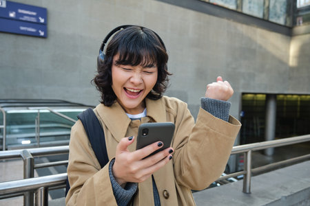 Happy asian girl in headphones, looking at mobile phone and celebrating, reading message with good...