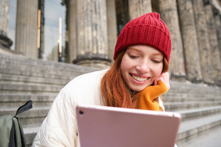 Portrait of young redhead woman sitting outdoors on stairs, reading e-book on digital tablet,...
