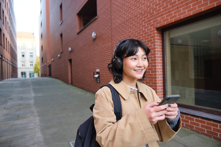 Beautiful smiling woman walking around town, listening music and using mobile phone app, going to...