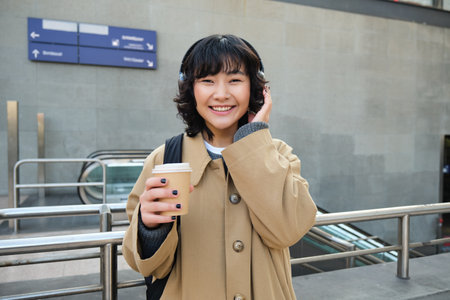 Beautiful smiling Korean girl, drinks takeaway coffee, listens music in headphones, wears trench and backpack, stands on train station, travellingの写真素材