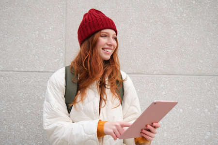 Young redhead woman with red hat, uses her digital tablet outdoors, stands on street with gadget, connects to wifi internet and searches for a location in internetの写真素材