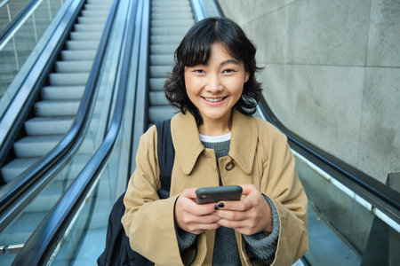 Portrait of beautiful asian girl, student goes down escalator in city, looks at mobile phone, uses telephone, map application, commutes somewhere in townの写真素材
