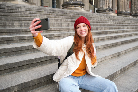 Stylish young girl in red hat, takes photos on smartphone camera, makes selfie as she sits on stairs near museum, posing for photo with app filterの写真素材