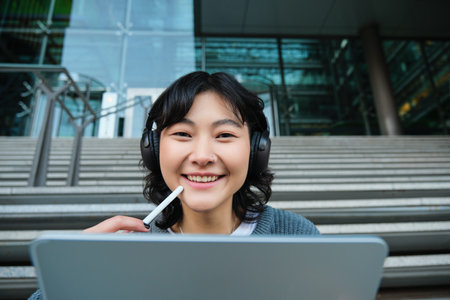 Close up of smiling asian girl feels inspired while draws on digital tablet, makes graphic design project, pencils user interface assignment, sits on stairs near campusの写真素材