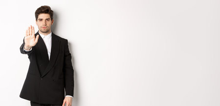 Portrait of serious handsome man in formal suit, extending hand to stop you, prohibit action, forbid and disagree with something, standing against white backgroundの写真素材