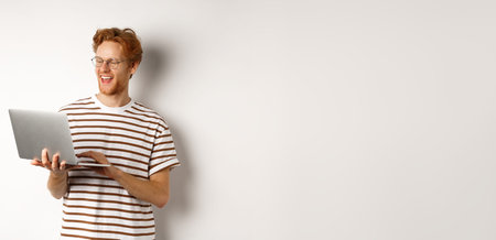 Handsome redhead man working freelance on laptop, smiling and looking pleased at computer screen,...