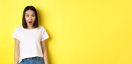 Beauty and fashion concept. Surprised asian girl open mouth and staring at camera, drop jaw impressed, standing over yellow backgroundの写真素材