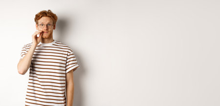 Young guy with ginger hair and glasses zipping mouth, showing lips seal gesture and smiling, keeping a secret, standing over white backgroundの写真素材